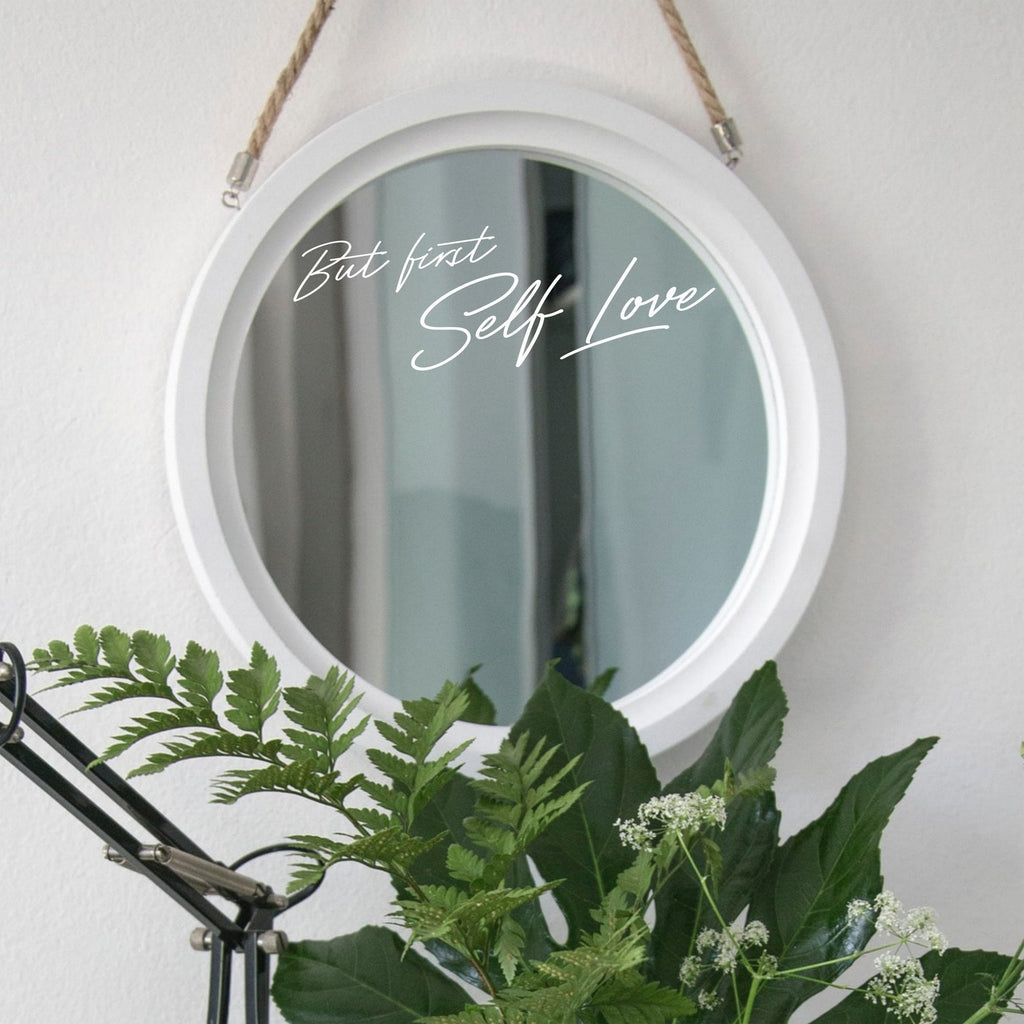 White “But First Self Love” mirror decal on a round white-framed mirror with green plants