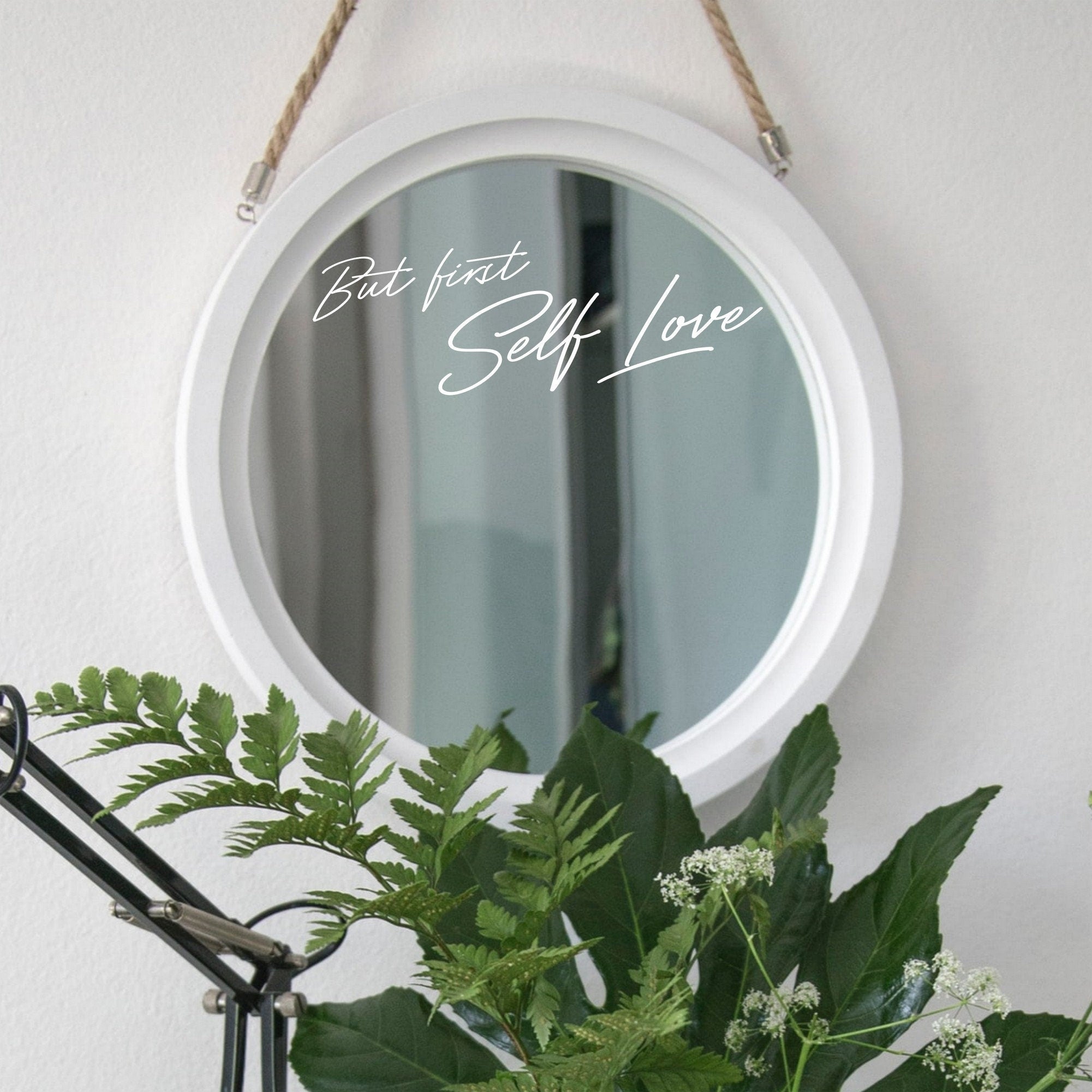 White “But First Self Love” mirror decal on a round white-framed mirror with green plants