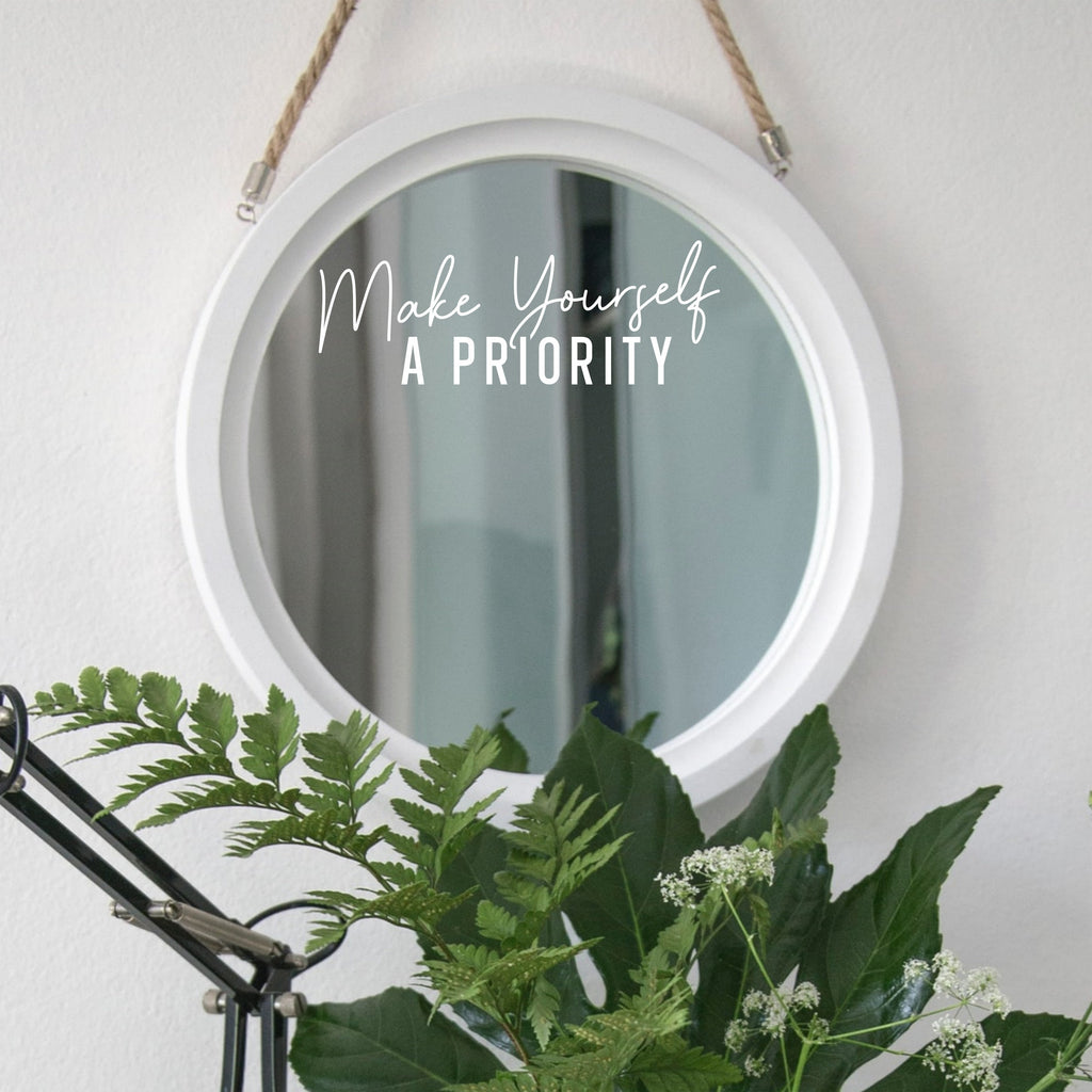 White “Make Yourself A Priority” mirror decal on a round white-framed mirror with green plants