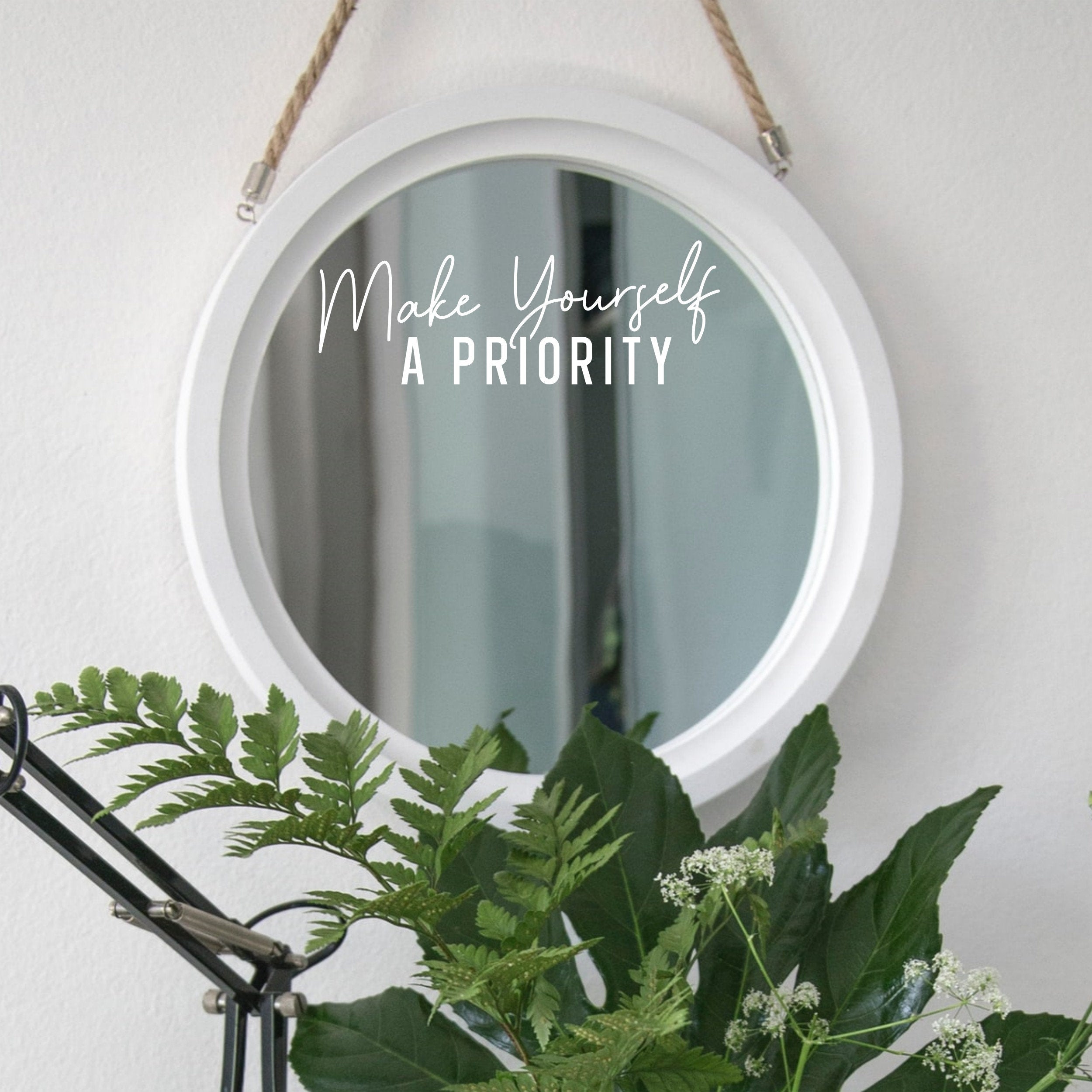 White “Make Yourself A Priority” mirror decal on a round white-framed mirror with green plants