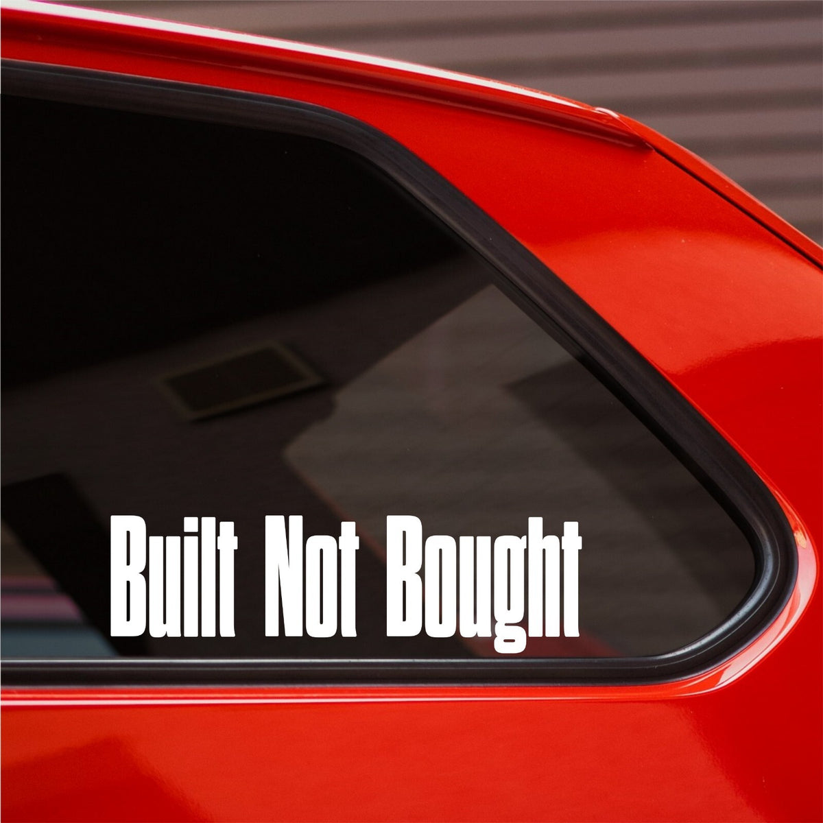 White Built Not Bought tall lettering decal on the rear side window of a red car.
